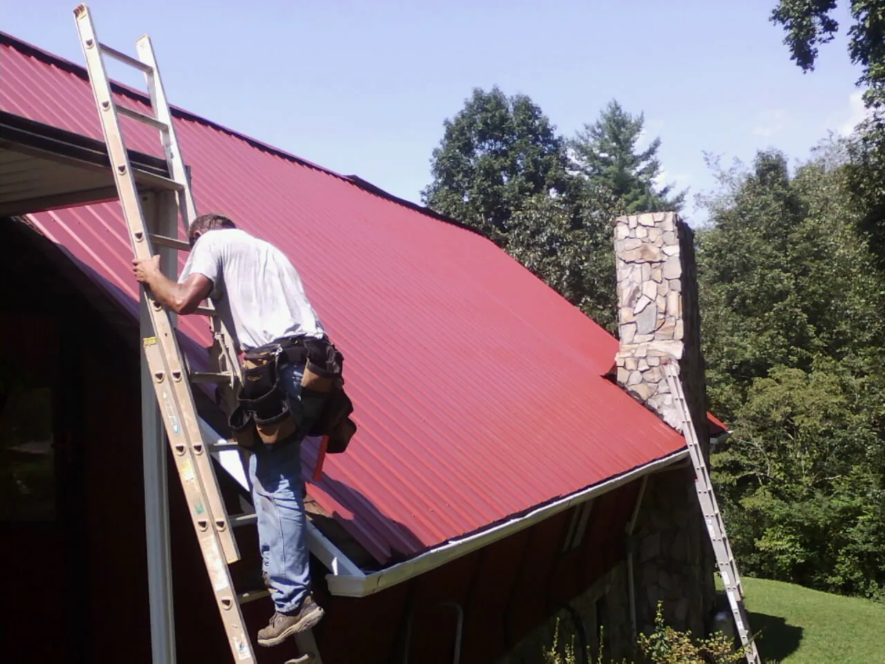 Roofer on a ladder beside a red metal roof and stone chimney in Shawnee